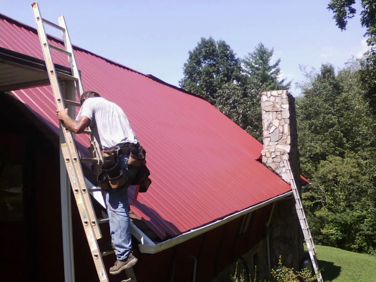 Roofer on a ladder beside a red metal roof and stone chimney — Soffit & Fascia Repair by Roofing Brothers in Woody Creek